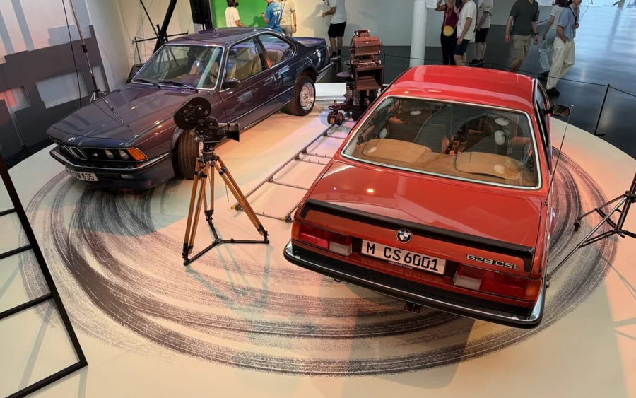 BMW Série 6 (E24) bleue et rouge exposées au Musée BMW entourées de visiteurs et de matériel de tournage
